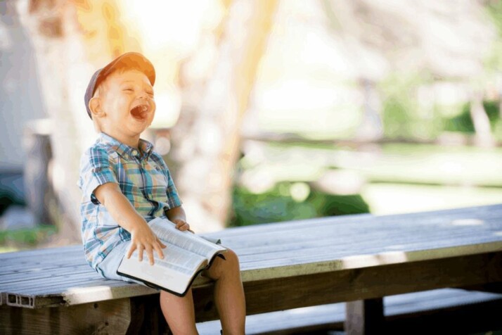 boy sitting on bench while holding a book ang laughing.