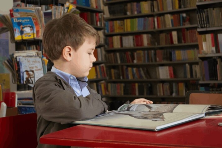 boy sitting near a red table and reading a book