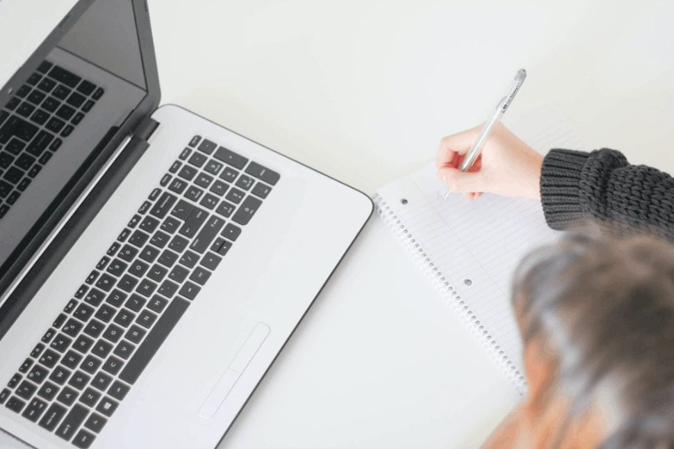 laptop on the table, woman sitting and writing with a pen in a book