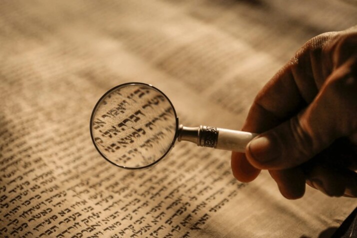 person holding a magnifying glass over a Hebrew manuscript.