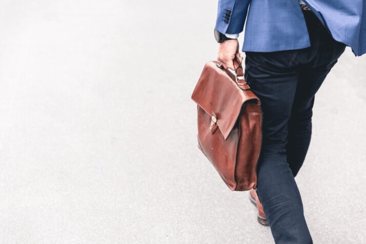 a man with a blue jacket walking holding brown leather bag.