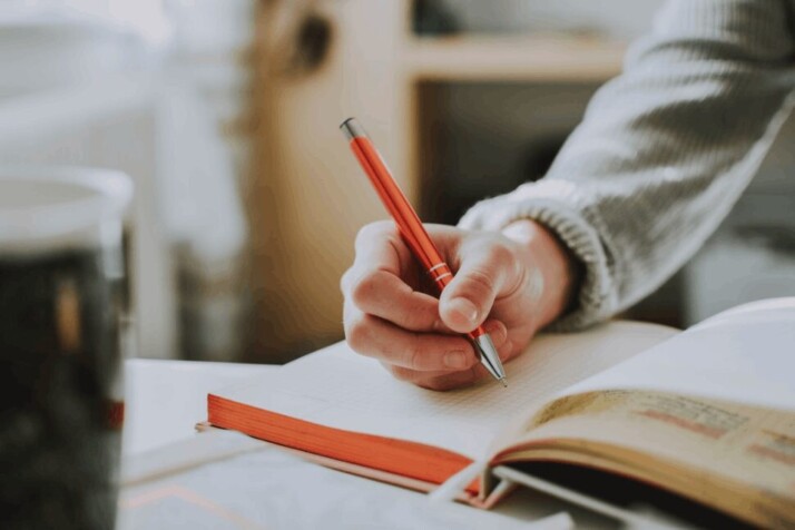 A person holding on red pen while writing on book.