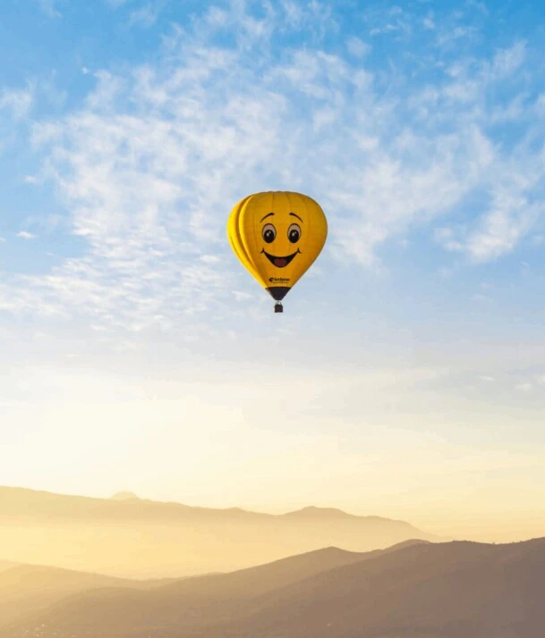 A smiling hot air balloon soaring mid air above a picturesque terrain.