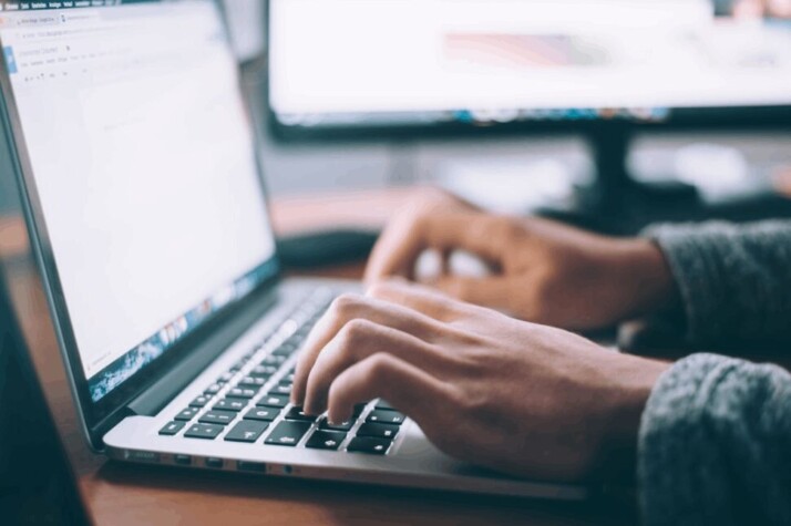 Laptop on a wooden table with hands typing on the keyboard.