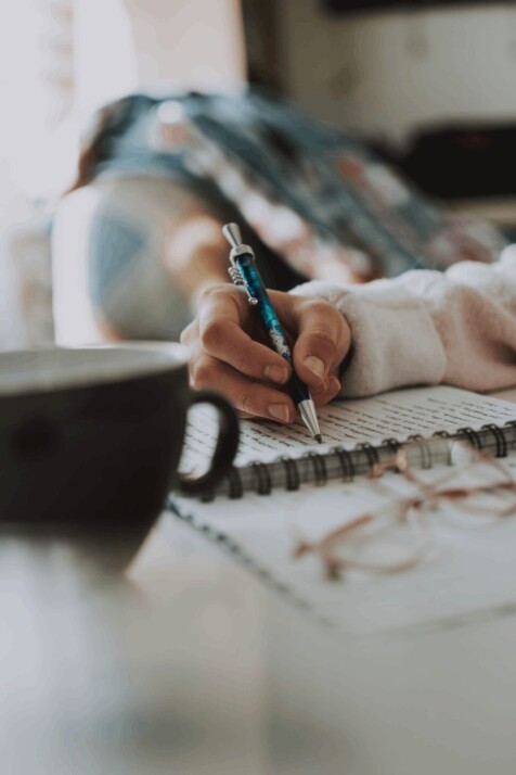 book, glasses and cup on table with person writing with pen on book.