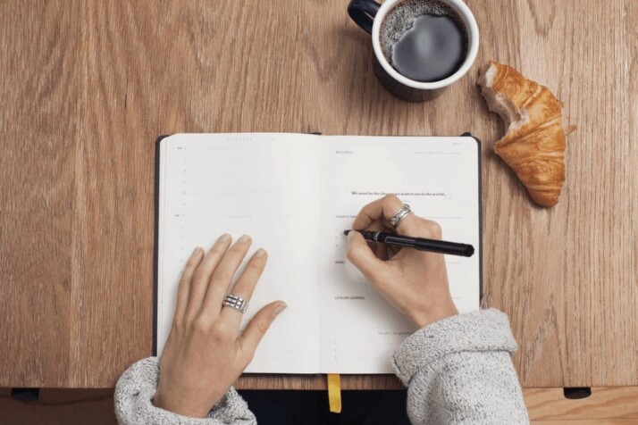 Person writing on a book with a cup of coffee and croissant on the table. 