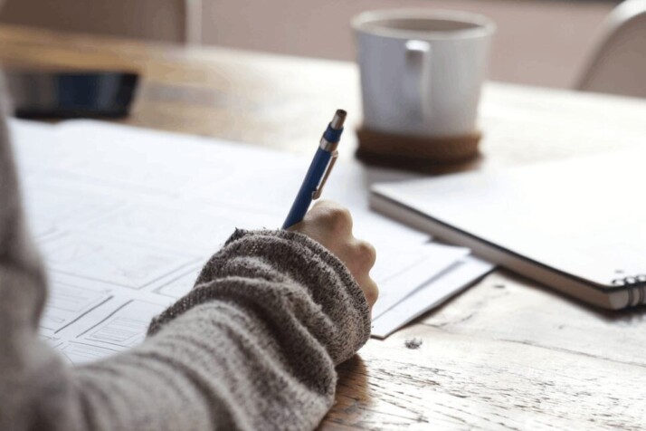 person writing on a brown wooden table near a white ceramic mug