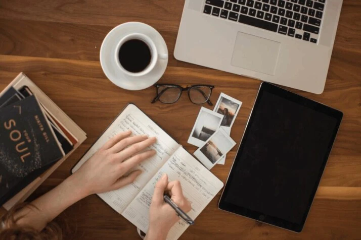 person holding ballpoint pen writing on notebook beside laptop