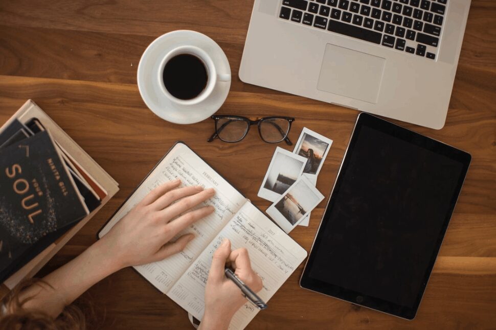 person holding ballpoint pen writing on notebook beside laptop