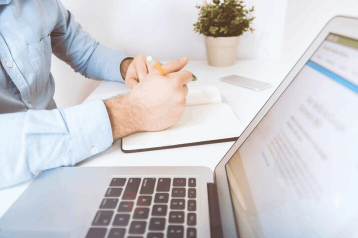 A person writing on white notebook next to a laptop