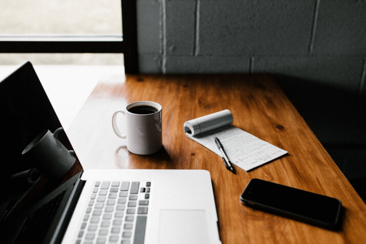 A laptop with a notebook, notepad, and phone on a wooden table. 