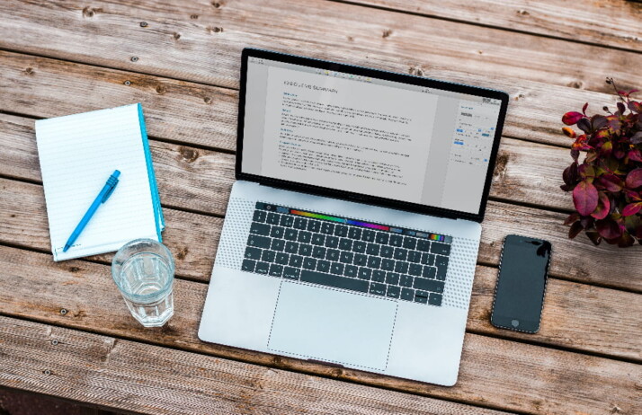 A laptop next to an iPhone, a notebook, and a glass of water.