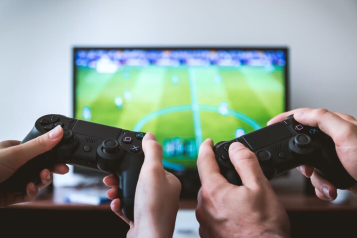 television in front with two people holding Black Gaming Consoles 