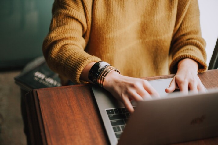 Person in an orange sweater typing on a Macbook.