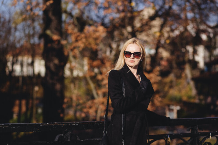 A blonde lady in a black overcoat and sunglasses on. 
