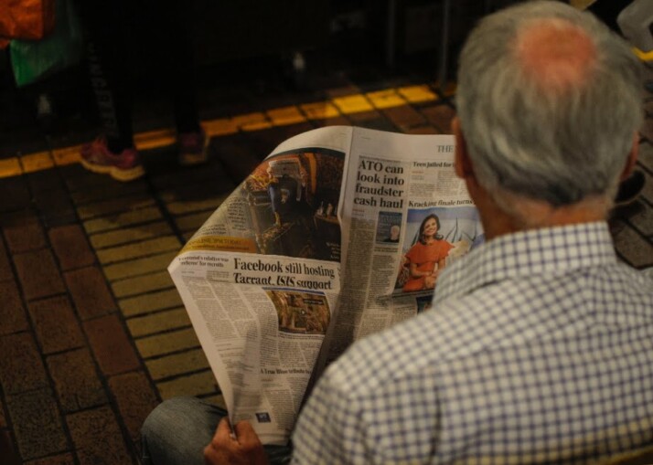 a slightly balding elderly man wearing a shirt reading a newspaper.