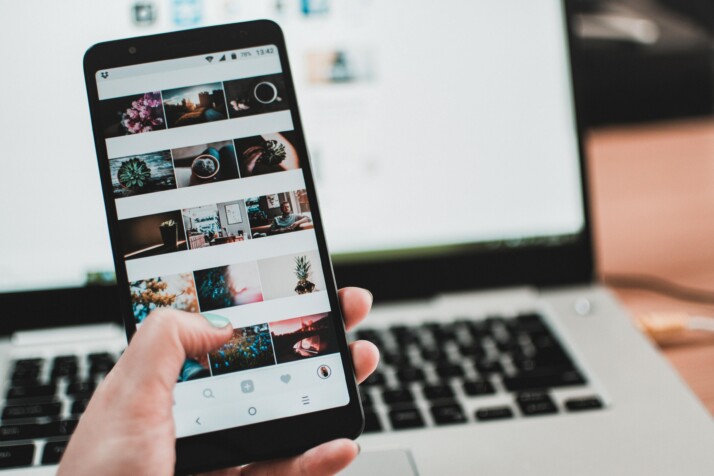 hand holding black phone displaying pictures with laptop in background