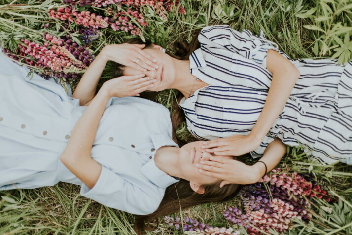 Two sisters holding each other's eyes closed while lying on a flower field.