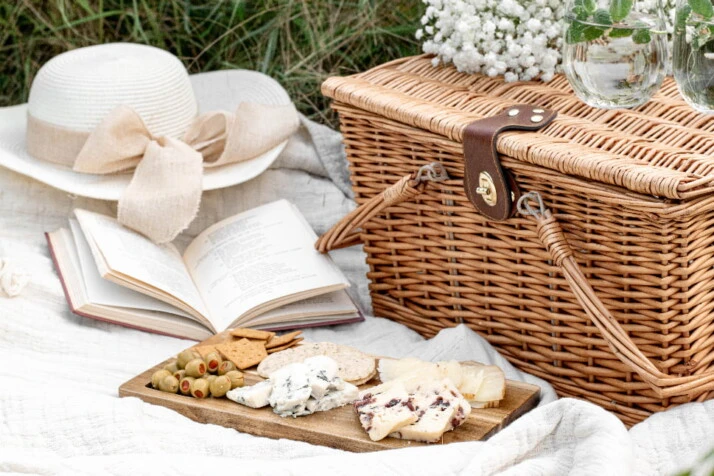 A beautiful picnic set-up with a book, hat, picnic basket, and a cheese board.