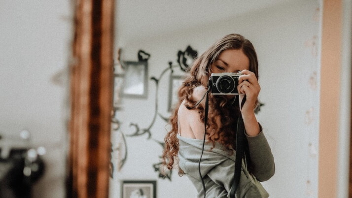 A mirror selfie of a woman in gray jacket holding black camera