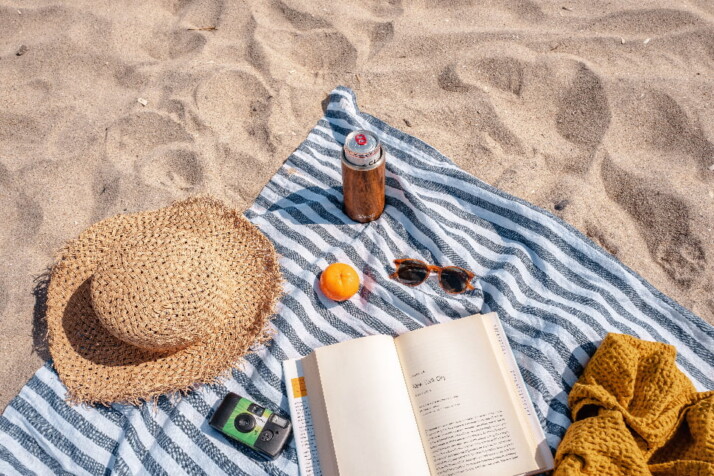 A book, camera, hat, sunglasses, and an orange placed on a blanket in the beach.