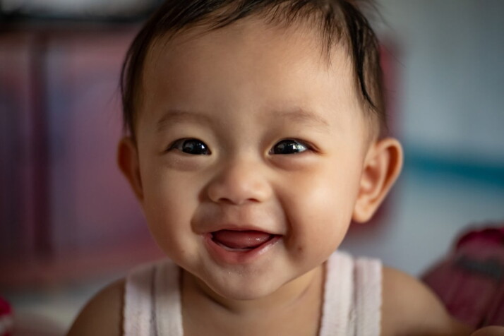 A portrait photo of a smiling baby wearing a white tank top. 