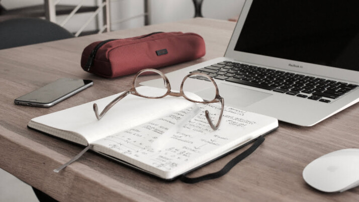 A MacBook Air next to an open notebook, glasses, a phone, and a pencil case.