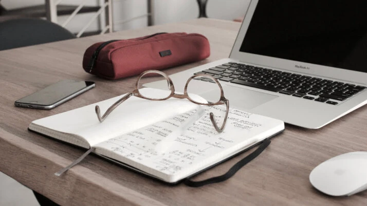A MacBook Air next to an open notebook, glasses, a phone, and a pencil case.