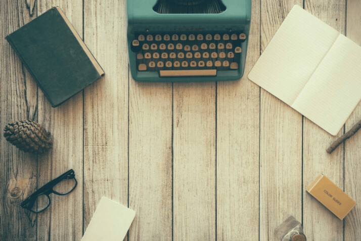 vintage teal typewriter beside book, eyeglasses, paper, and pencil