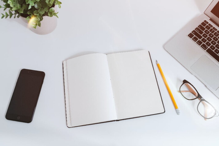 A blank notebook next to a pencil, iPhone, and reading glasses.