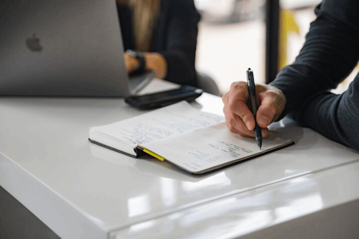 A person writing on a white notebook placed on a table. 