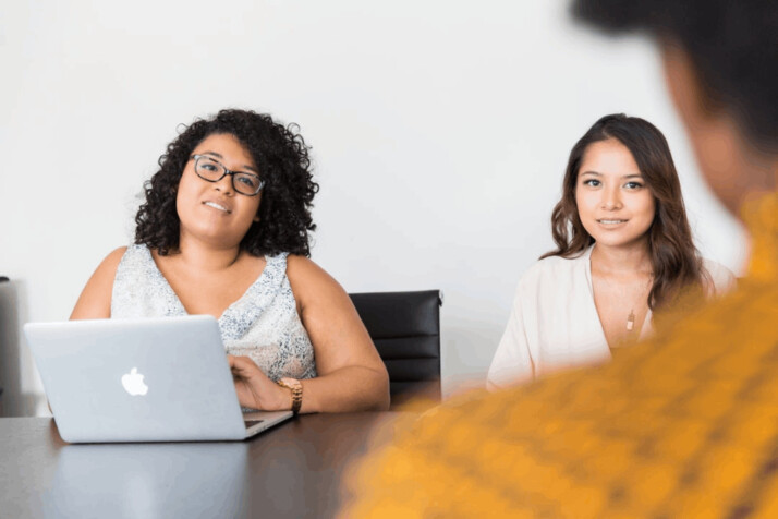 two women with a MacBook looking at person across the table