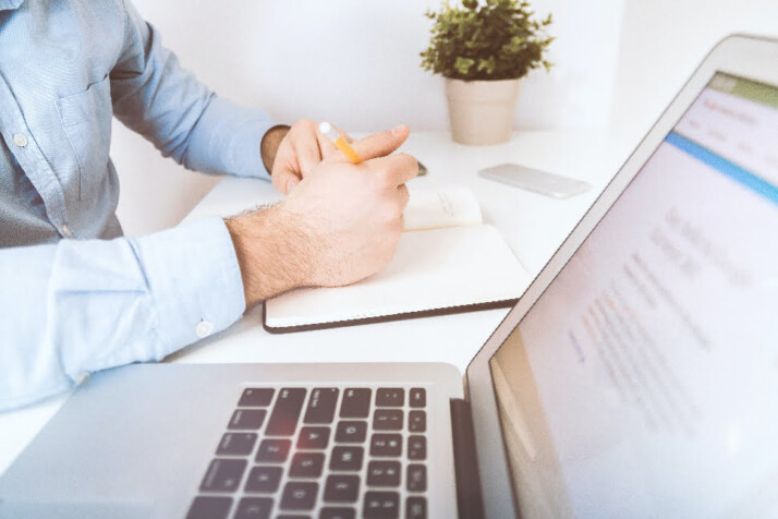 A person writing something down on a notebook, while looking at a laptop.
