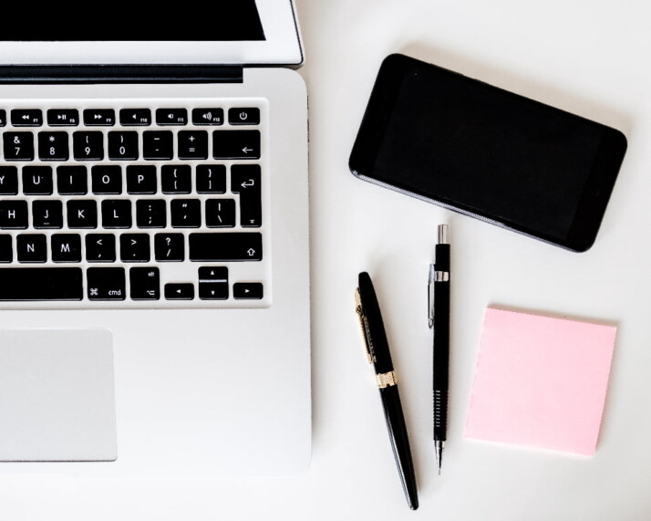 A laptop next to a phone, two pens, and a note pad.
