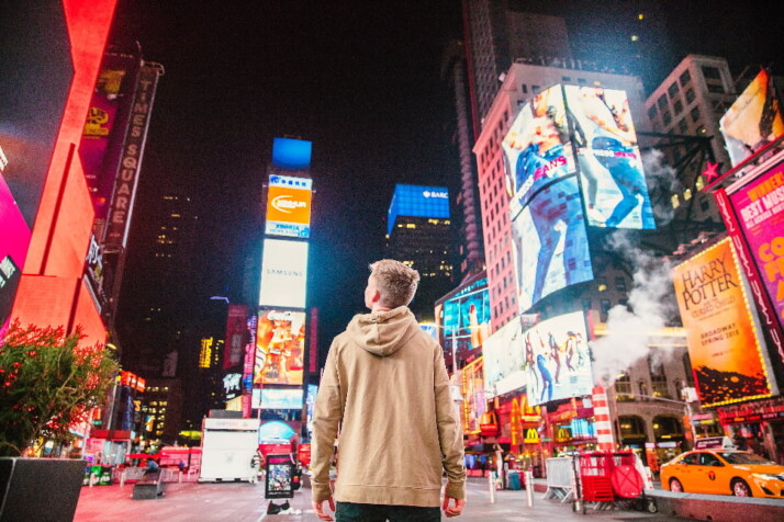 a blonde man in a beige sweatshirt in the middle of times square in night.