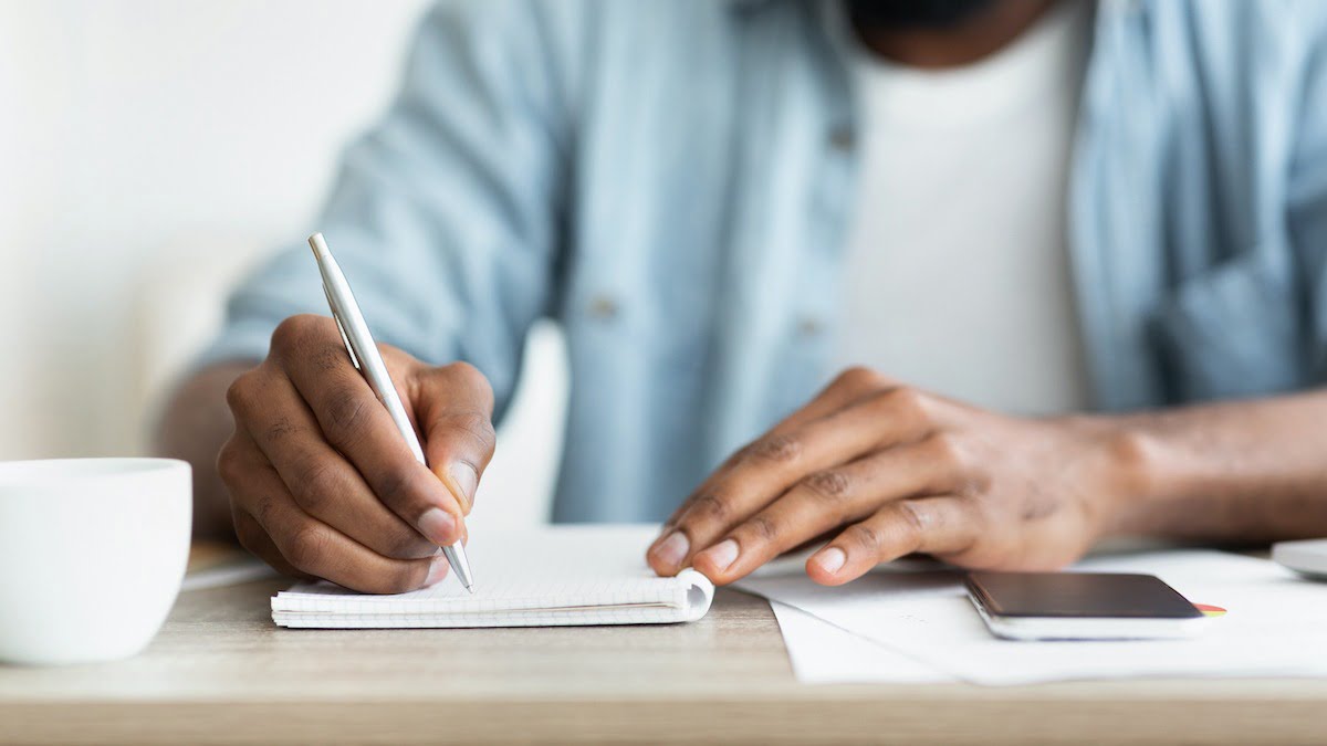 man wearing blue shirt writing in book with white pen