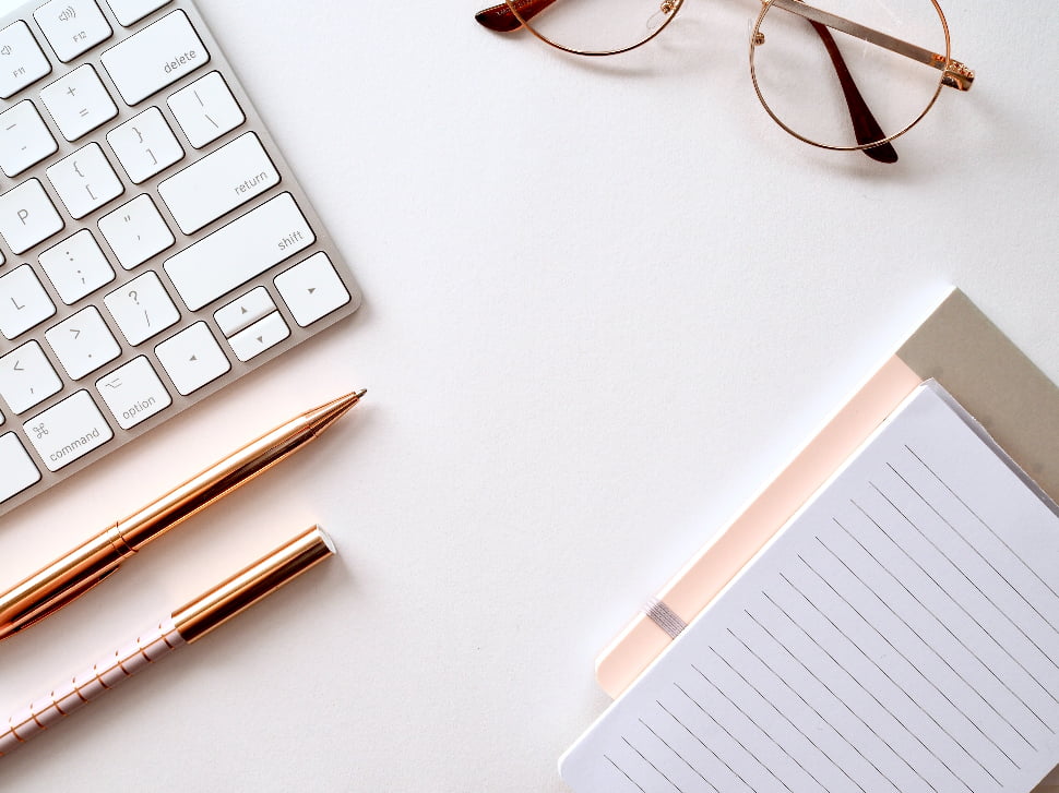 A notebook, reading glasses, pens, and a keyboard placed next to each other.