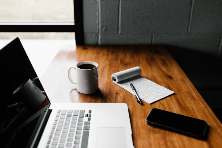 A laptop, a cup of coffee, a phone, and a jotter on a brown surface