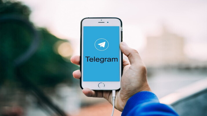 person wearing blue sweater holding white Iphone displaying telegram page