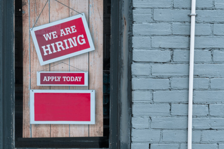 window in a brick wall that has placards which read we are hiring apply today.