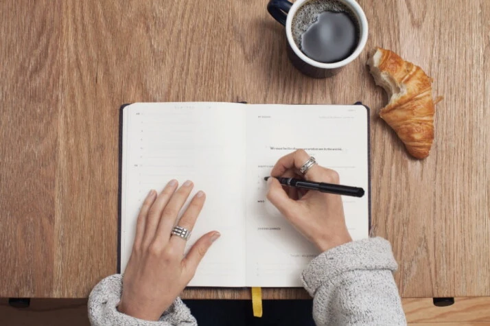 A person writing on a notebook with a croissant and cup of coffee beside them.