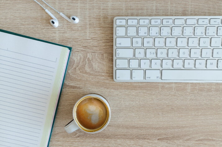 A notebook, keyboard, earphones, and cup of coffee all placed on a table.