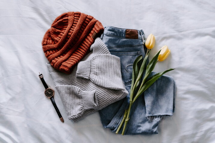 A sweater, cap, jeans, and watch is placed on the bed with two yellow tulips. 