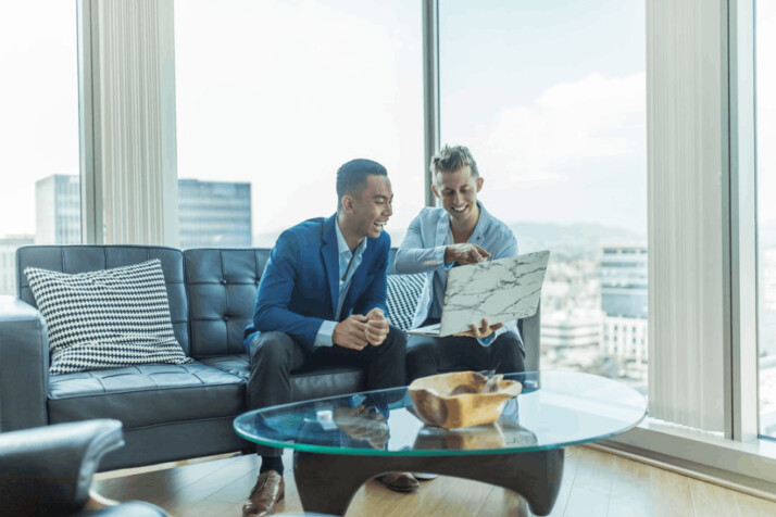 two men in suit sitting on sofa laughing and looking into laptop computer