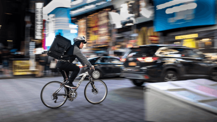 A man riding a bicycle near vehicles to deliver items.