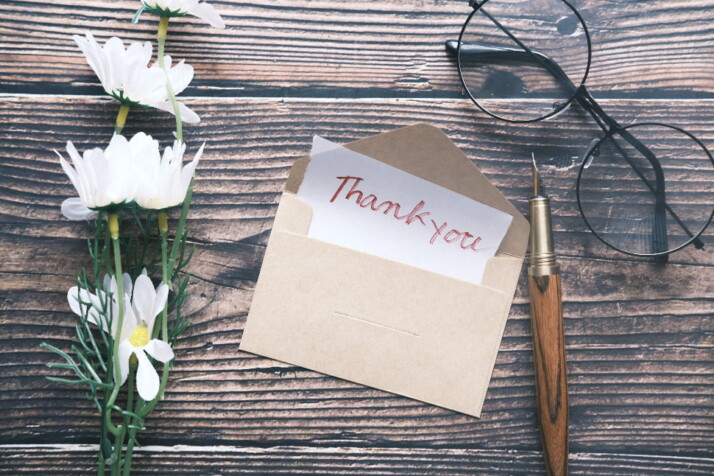 A thank you letter next to a pen, glasses, and white flowers.