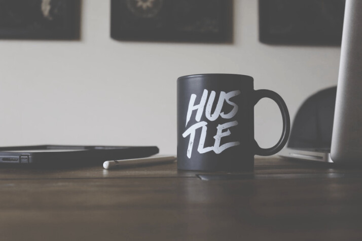 black and white Hustle-printed ceramic mug on table next to an open macbook