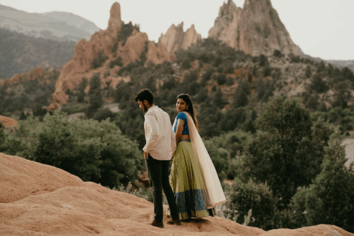 a brown ethnic couple standing over a rock, looking at mountains.