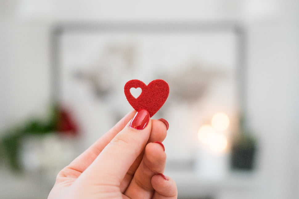 A red heart cut-out held by a hand with red nails.