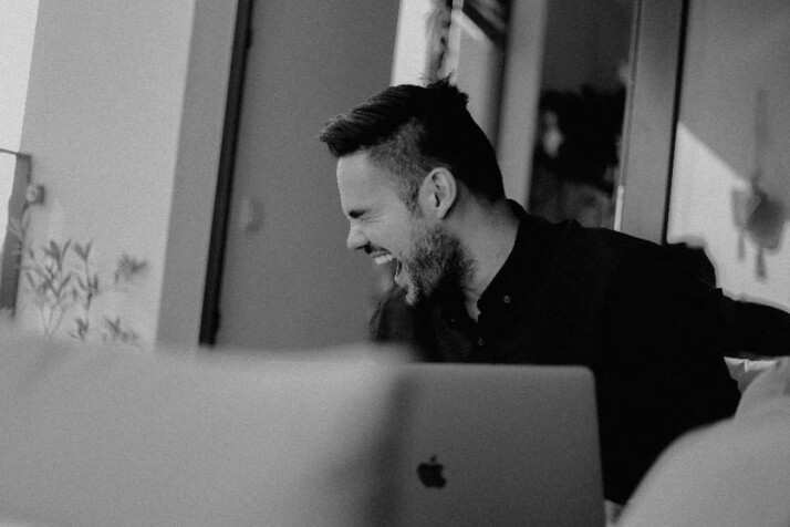 A black and white photo of a guy laughing in front of his laptop.